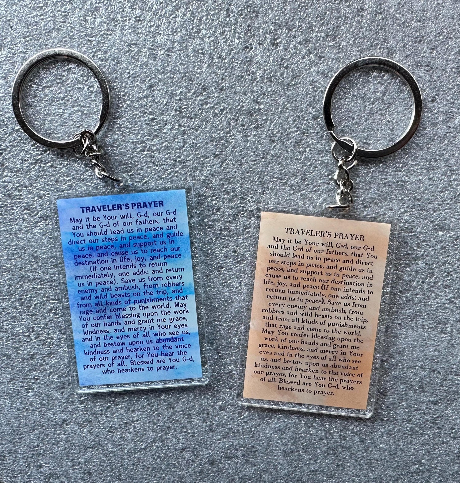 acrylic keychains with a blue watercolor background. Each keychain has a white rectangle with the text 'TRAVELER'S PRAYER' and a prayer written in black text. The prayer is the same on both keychains.