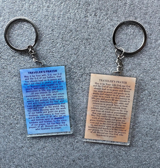 acrylic keychains with a blue watercolor background. Each keychain has a white rectangle with the text 'TRAVELER'S PRAYER' and a prayer written in black text. The prayer is the same on both keychains.